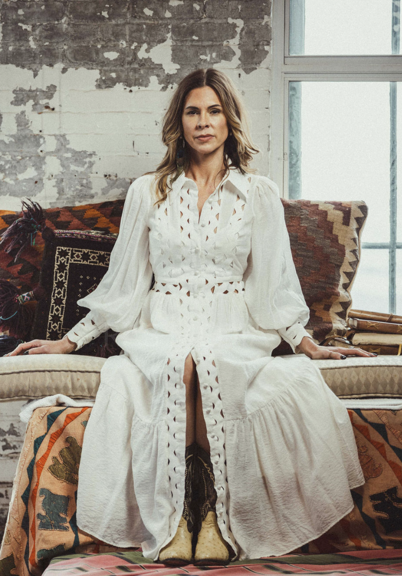 Misti Aaronson seated on a sofa with woven kilim pillows against a white brick wall